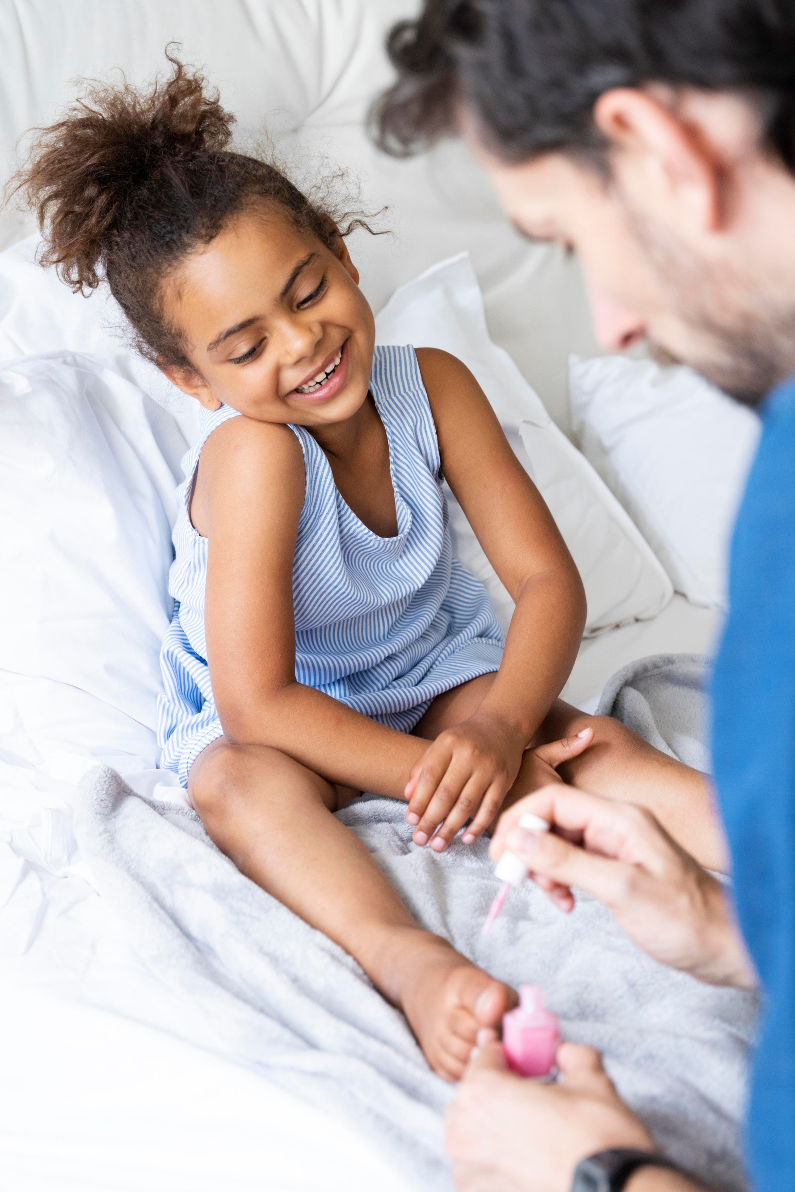 A parent painting a young child's toenails on a bed — a warm, candid family moment at home.